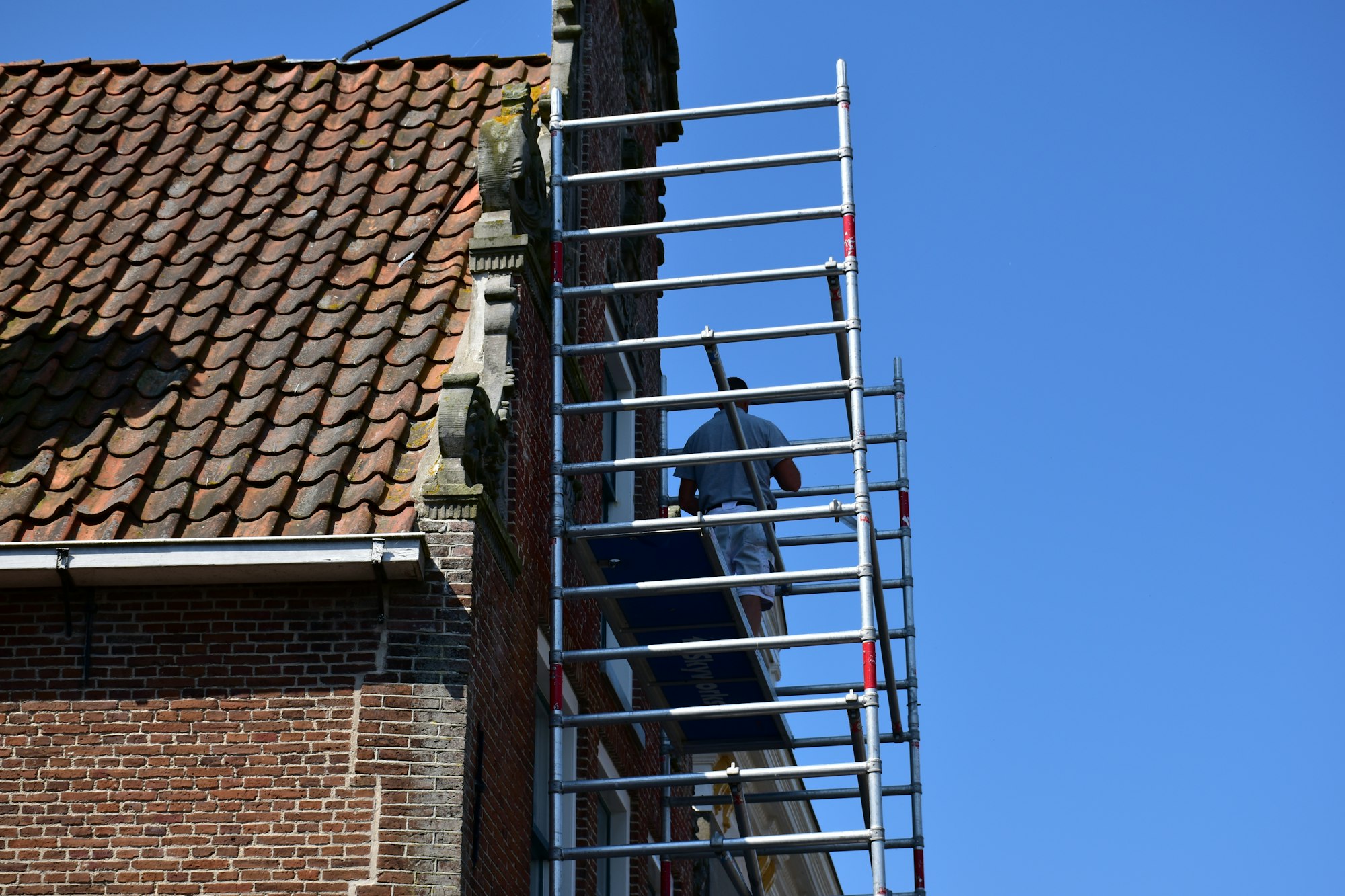 Painter on scaffolding coating the exterior of a brick building under a clear blue sky